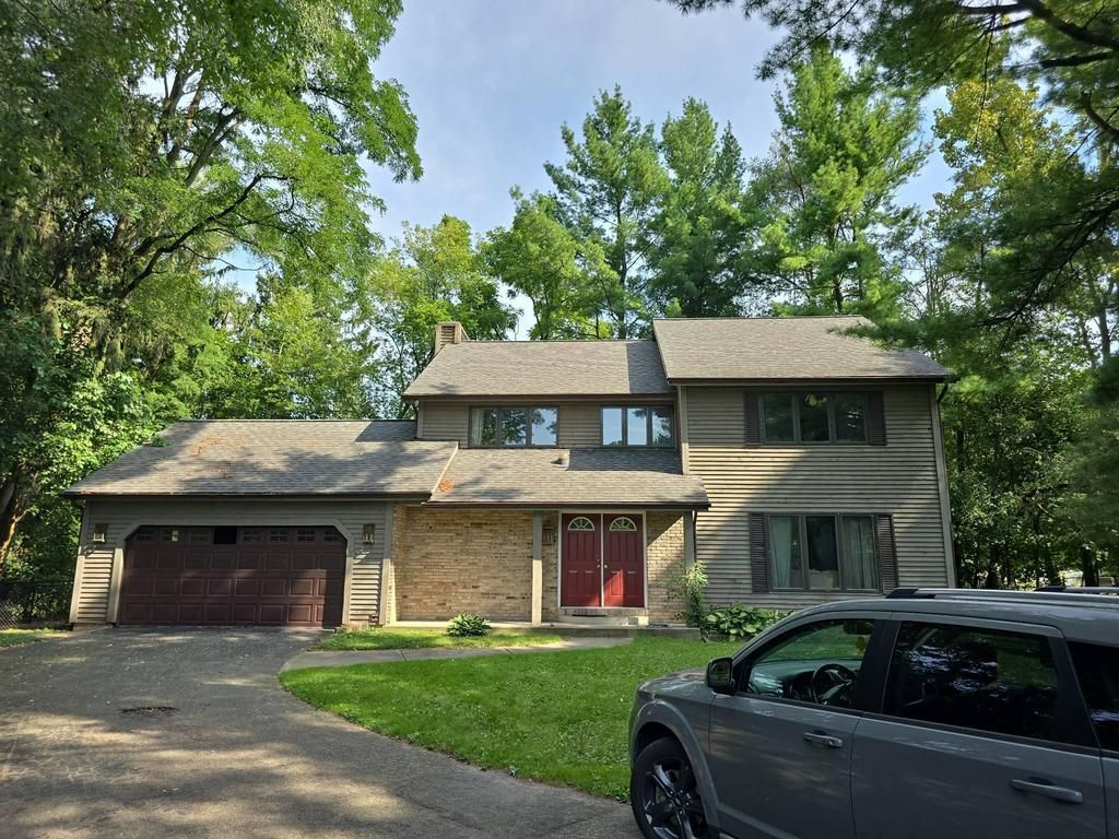 A two-story house with a brick and light-colored siding exterior, a garage, and a parked car, surrounded by mature trees.