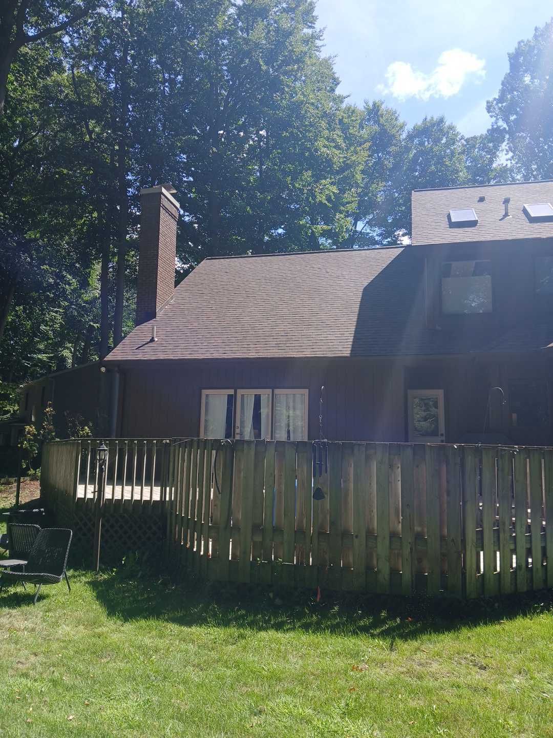 A brown two-story house with a wooden deck and a brick chimney, seen from a grassy yard on a sunny day.