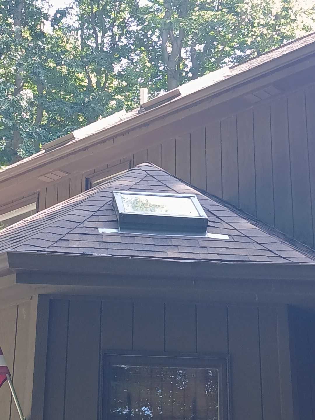 A skylight installed on a dark brown shingled roof section extending from a house with dark wood vertical siding.
