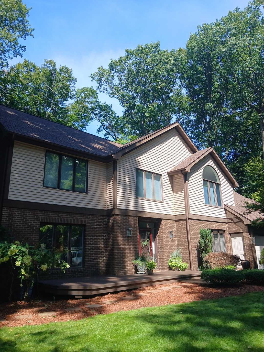 Two-story house with tan vinyl siding and brown brick lower level, surrounded by trees under a clear blue sky.