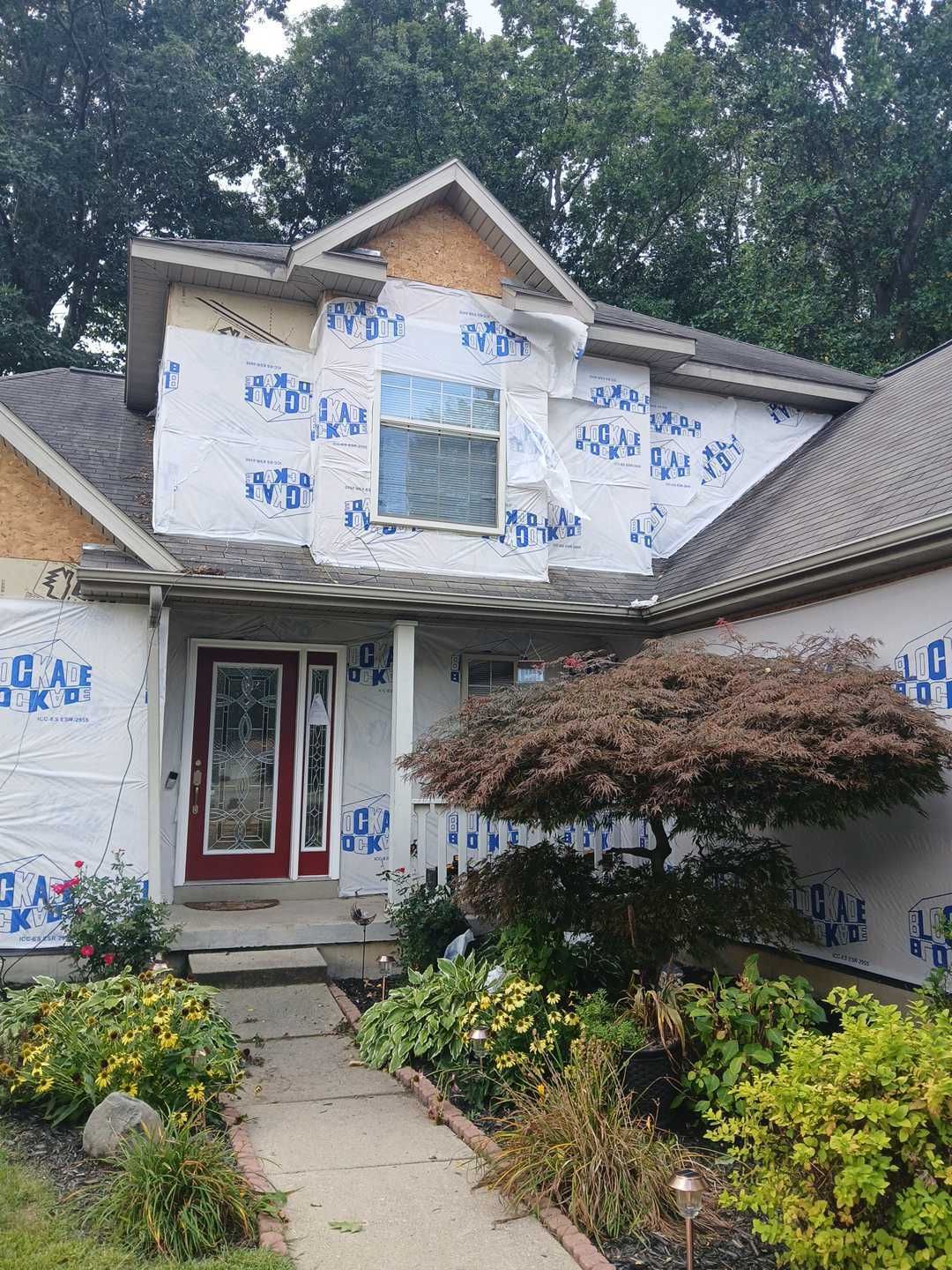 A two-story house exterior under construction, featuring white house wrap, a red front door, and a tree in the yard.
