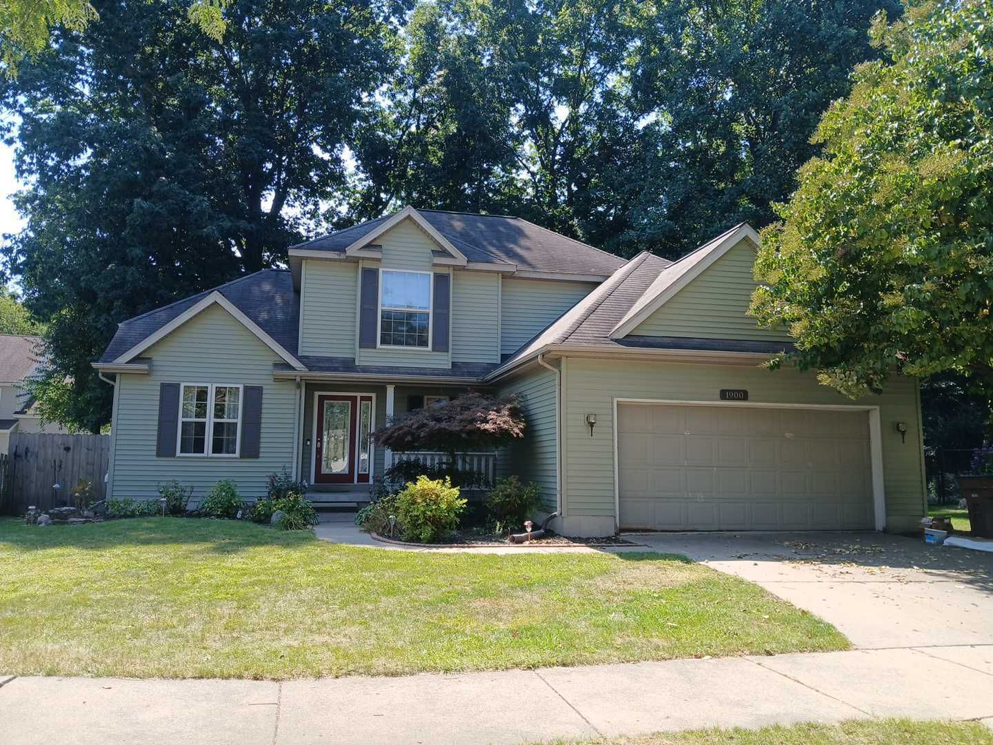 A two-story, sage-green suburban house with a two-car garage, front lawn, and surrounding trees under a blue sky.