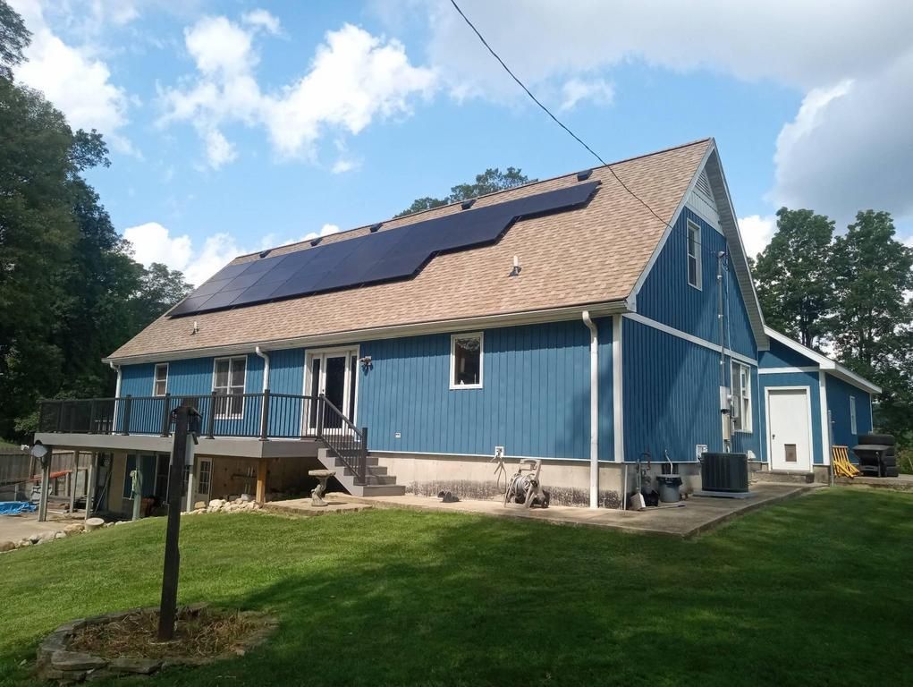 A blue house with solar panels on its roof, a wooden deck, and a grassy backyard under a partly cloudy sky.
