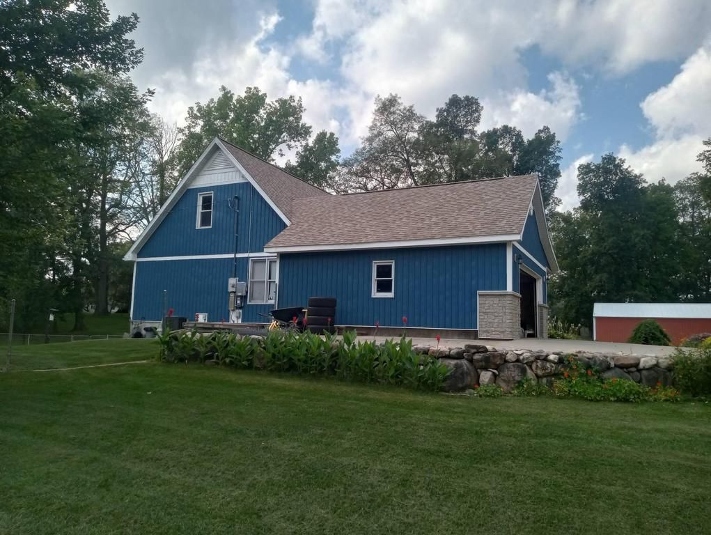 A blue house with a light brown shingled roof, surrounded by green trees and a lawn with a low rock retaining wall.