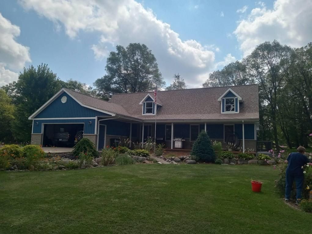 A blue, single-story house with a garage, dormer windows, and a brown shingle roof, set in a sunny, tree-lined yard.