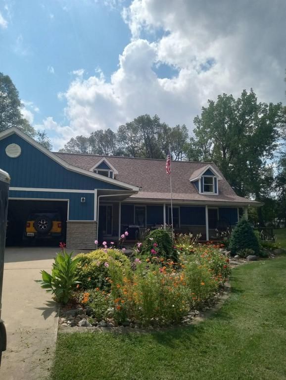 Blue house with a stone foundation, garage, and flower garden, set against a tree-lined backdrop under a cloudy sky.