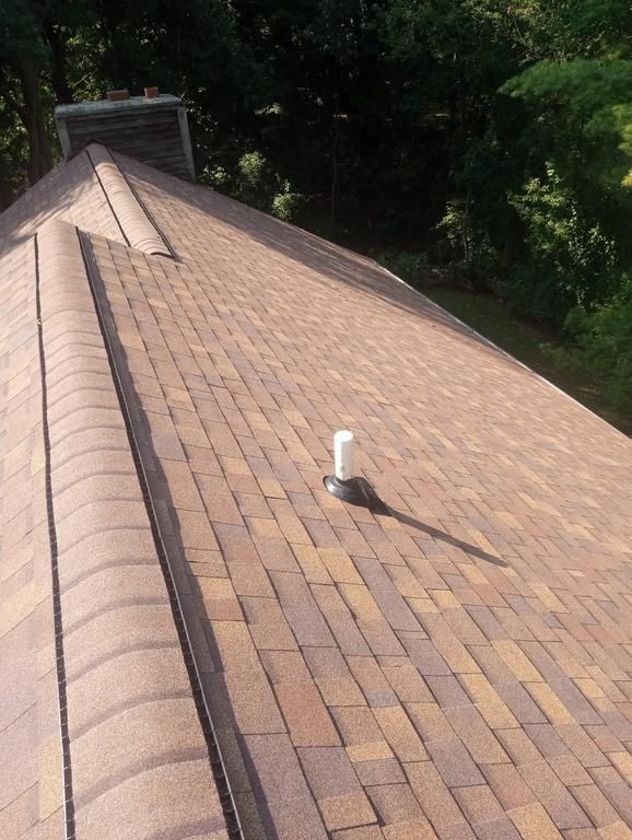 A brown asphalt shingle roof on a sunny day with a white pipe vent and a brick chimney in the background.