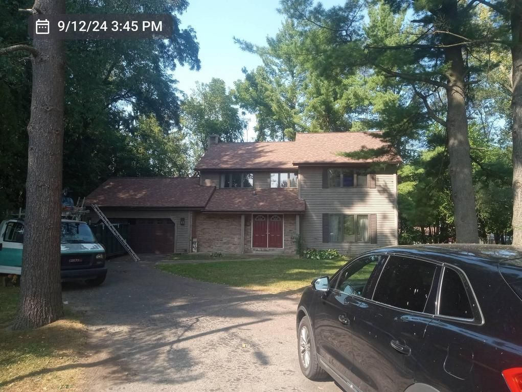 Two-story house with a brown shingled roof, red front doors, and brick siding, nestled among tall trees with a driveway.