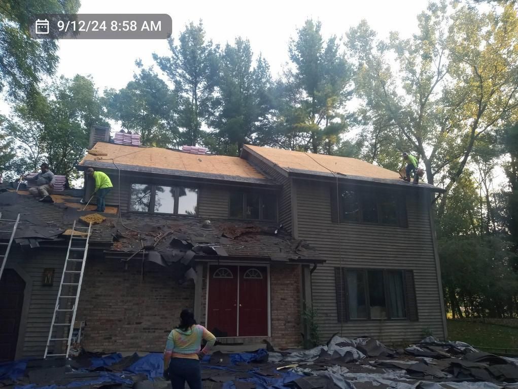 Workers are stripping old shingles from the roof of a two-story home, leaving parts of the wooden decking exposed.