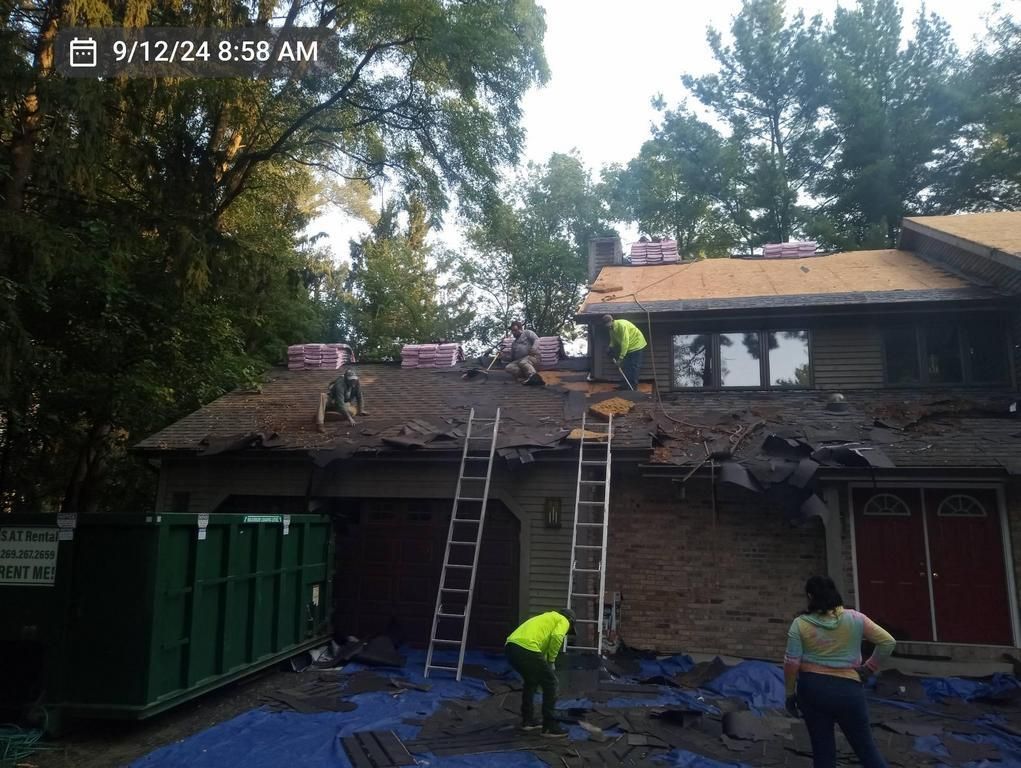 Construction crew members working on a residential roof replacement with a green dumpster parked in the front yard.
