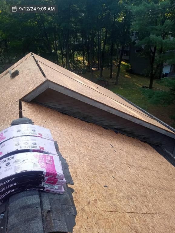 A view from a roof under construction, showing plywood sheathing and stacked roofing shingles with trees in the background.