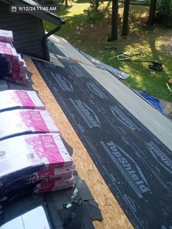 Stacks of pink-wrapped roofing shingles on a residential roof, with black underlayment partially installed on the deck.