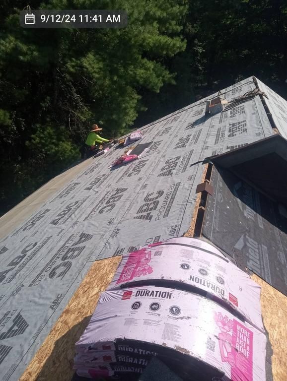 A roofer works on a sloped roof covered in underlayment, with bundles of shingles in the foreground.