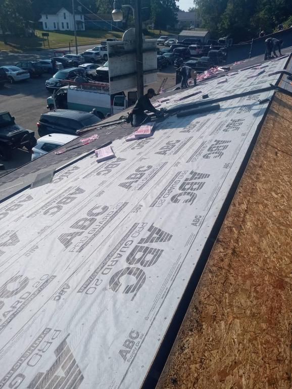 Construction workers install synthetic roofing underlayment on a steep, slanted wooden roof.