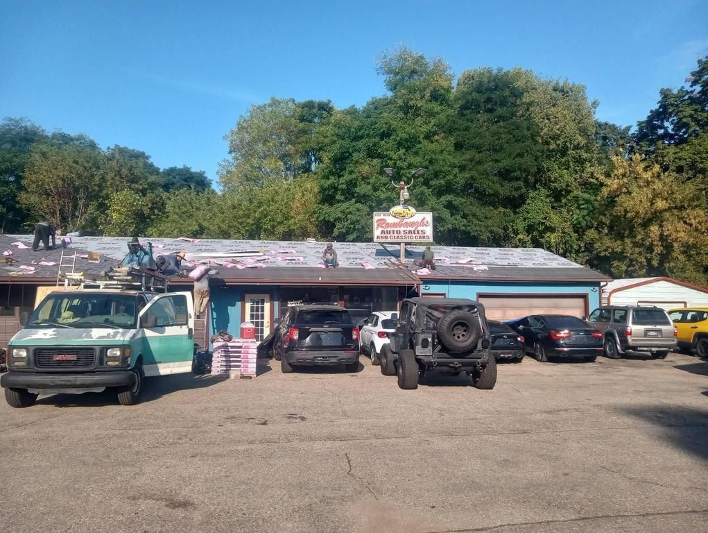 Workers repair the roof of a blue commercial building with several cars and a utility van parked in front.