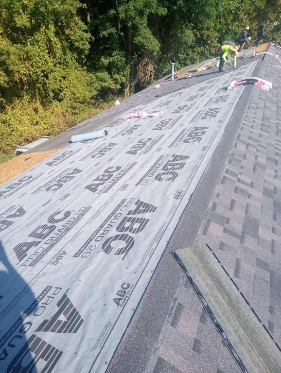 Workers install new shingles and protective underlayment on a sloping roof, with trees in the background.