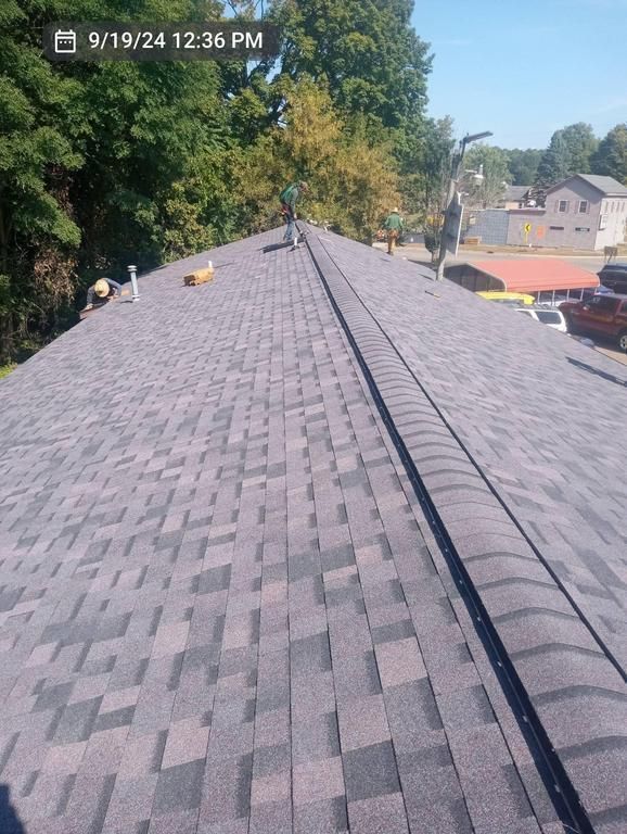 View from the roof ridge of a newly installed shingle roof, with a person working in the distance.