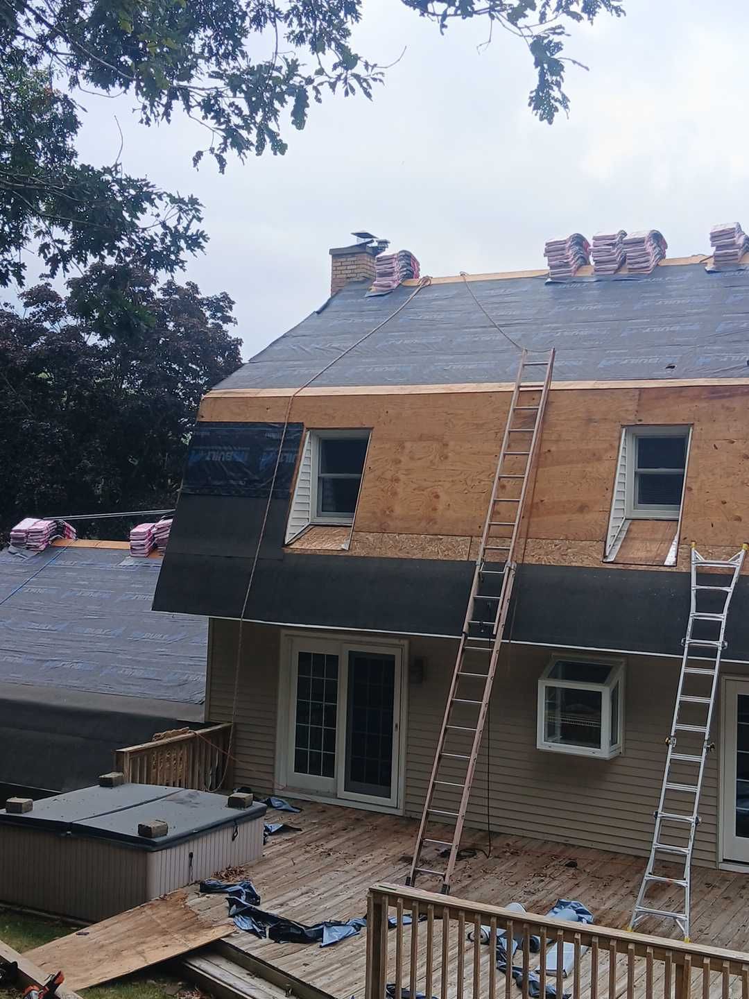 A house under construction with a partially shingled roof, exposed wooden siding, two ladders, and a deck.