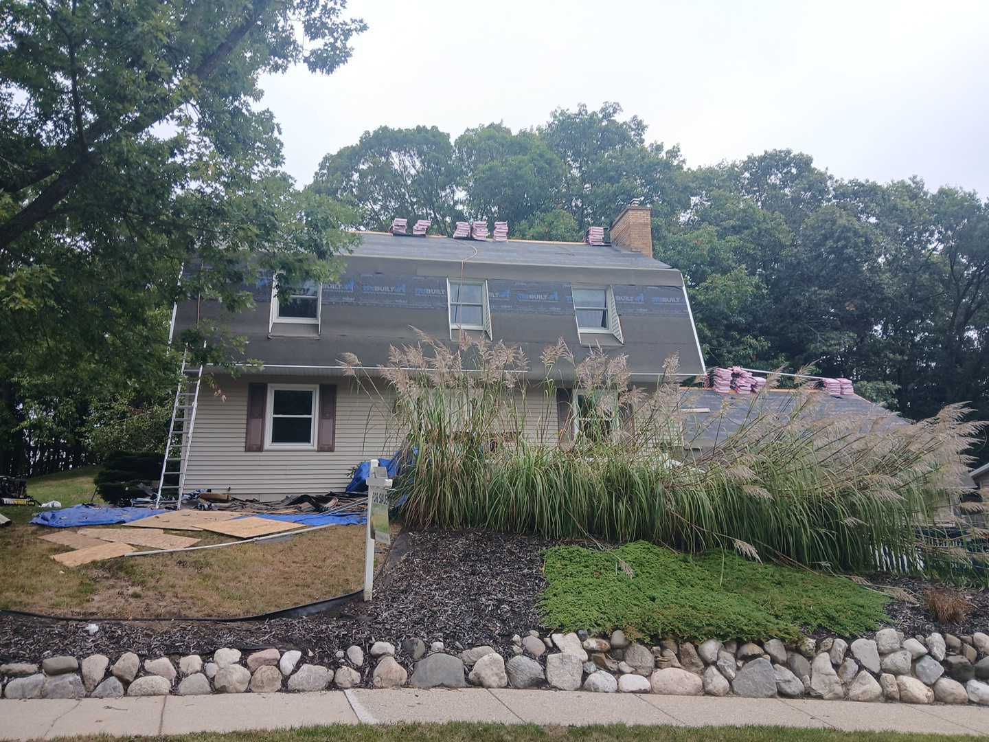 A house undergoing roof repairs, with workers visible on the roof, surrounded by trees and a stone retaining wall.