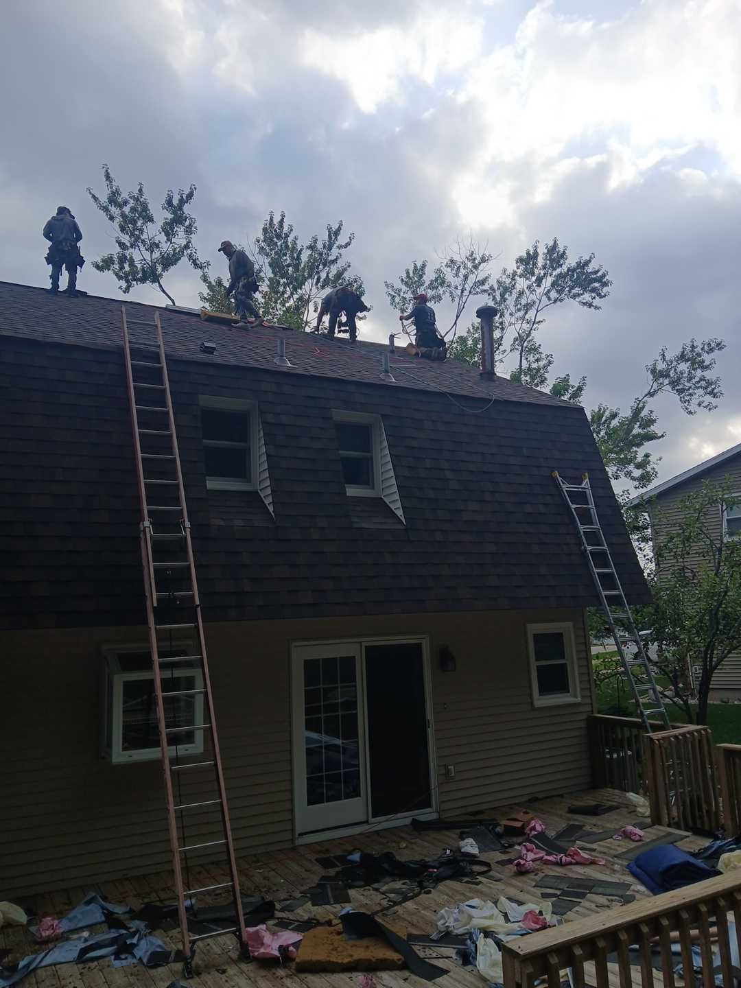 Four workers are on the steep, dark shingled roof of a house, with ladders and construction debris on the deck below.