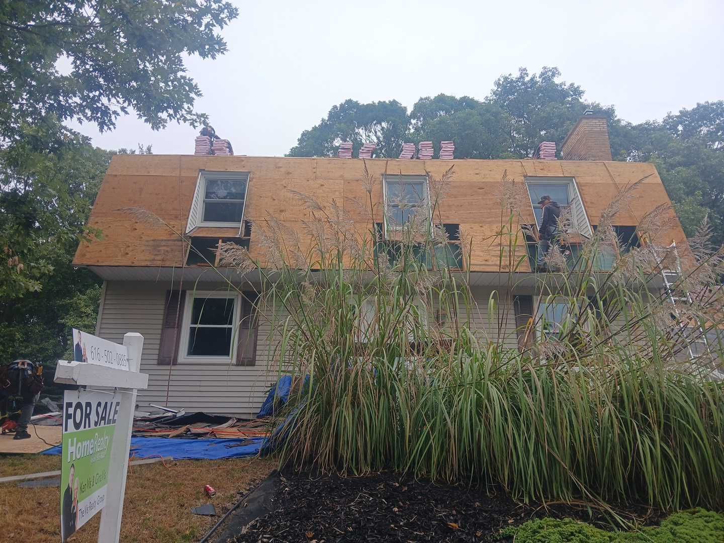 A house undergoing a roof replacement with exposed plywood sheathing, workers on the roof, and a for-sale sign in the yard.