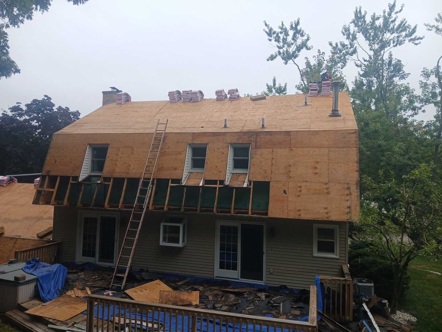 A partially re-roofed house with plywood sheathing, dormer windows, and a ladder leaning against the exterior.