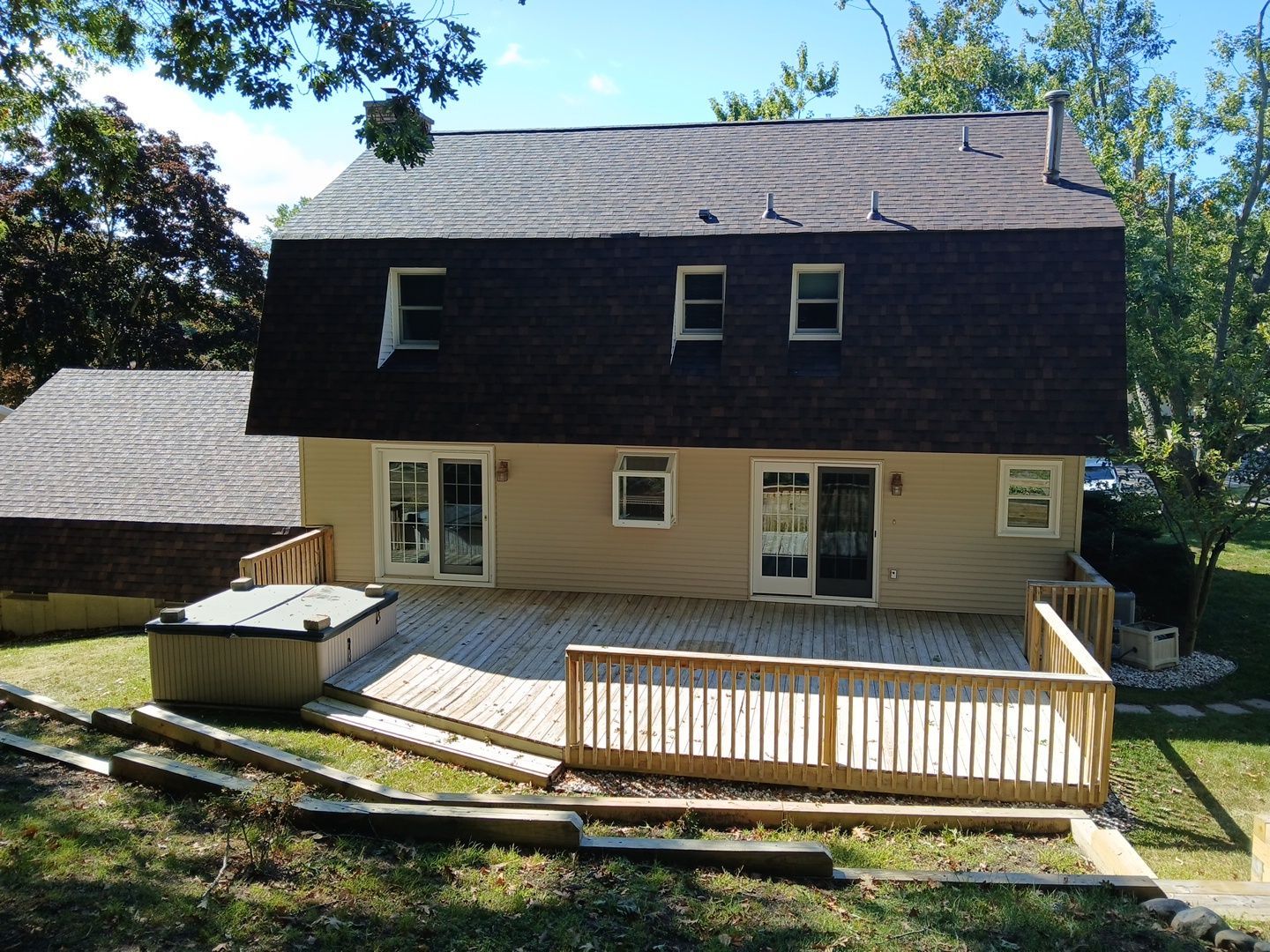 A beige house with a dark gambrel roof, featuring a new wooden deck and hot tub in the backyard.