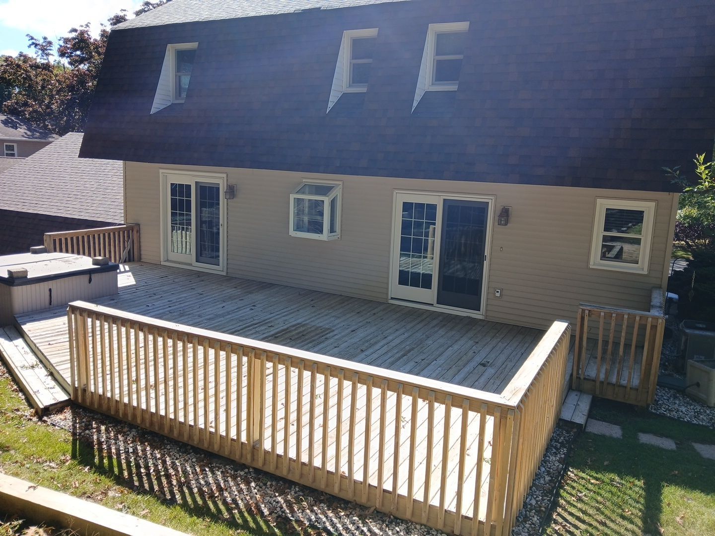 A wooden deck with a hot tub extends from the back of a tan house with a dark, sloped roof and several upper windows.