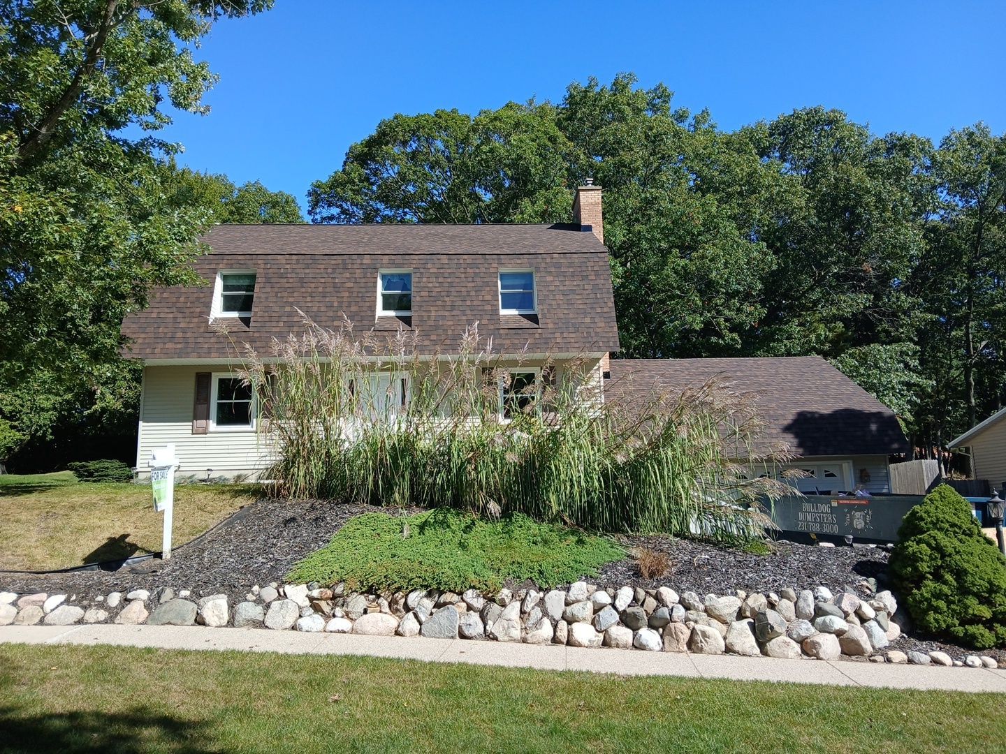 A two-story tan house with a brown shingled roof, set behind a rock retaining wall and green landscaping on a sunny day.