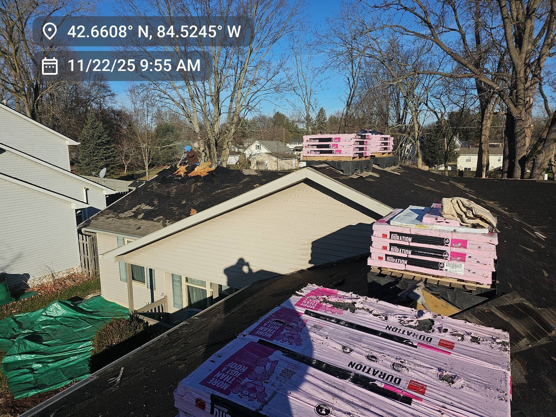 Bundles of pink roofing shingles sit on a residential rooftop under a clear blue sky, with a house visible nearby.