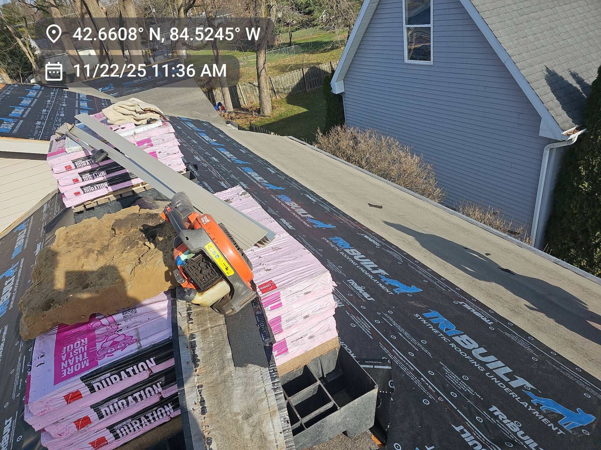 Stacks of pink roofing shingles and a power tool on a roof underlayment-covered slope next to a house.