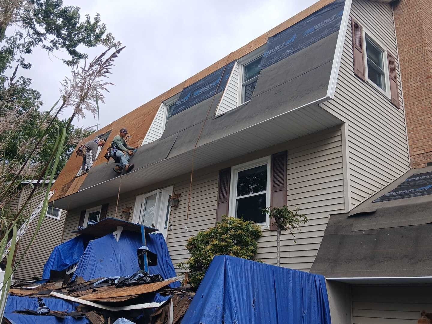 Two workers install roofing shingles on the steep mansard roof of a tan siding house, with blue tarps on the ground.