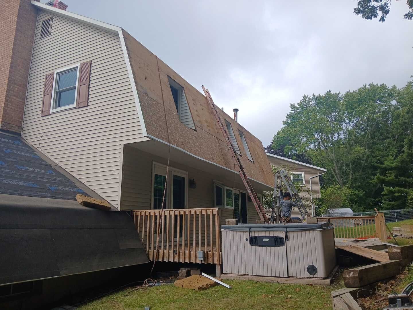 A worker uses a ladder against a house with a steep roof, next to a deck and a hot tub on a cloudy day.