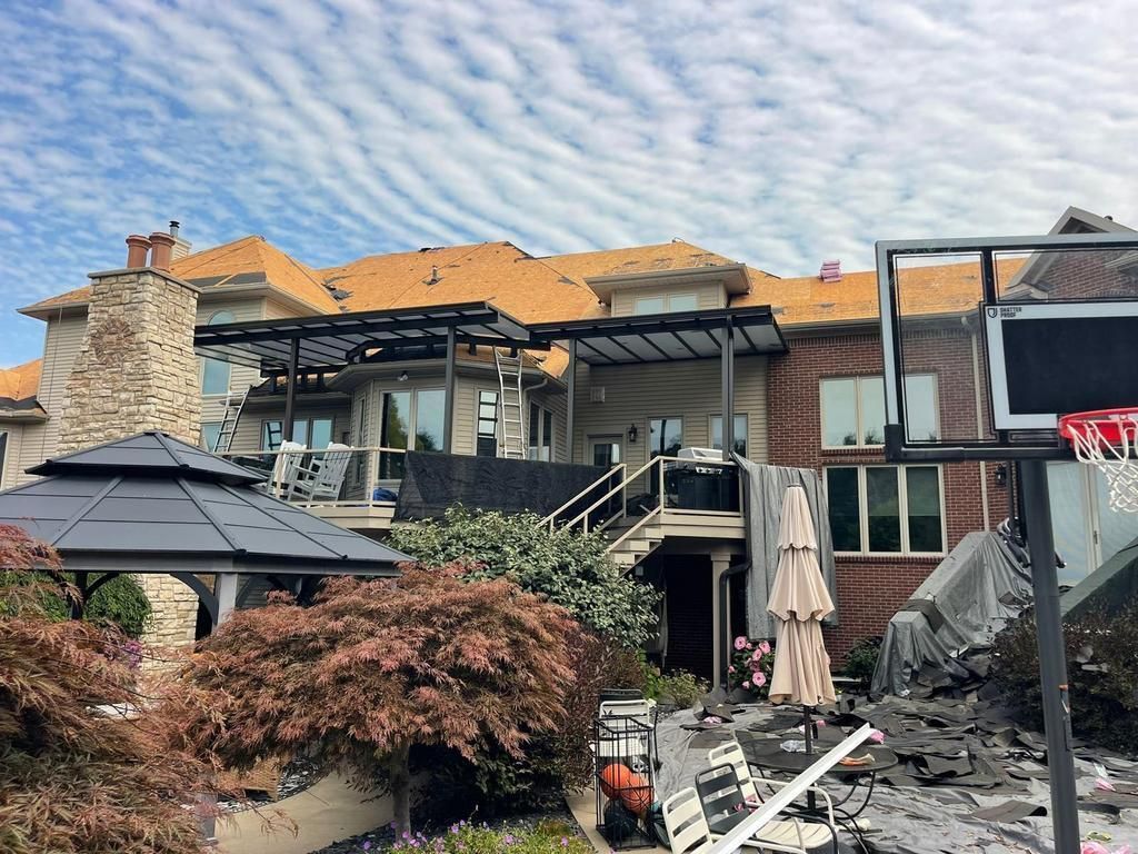 A large house exterior with a damaged roof under repair, a dark gazebo, and a basketball hoop in the backyard.