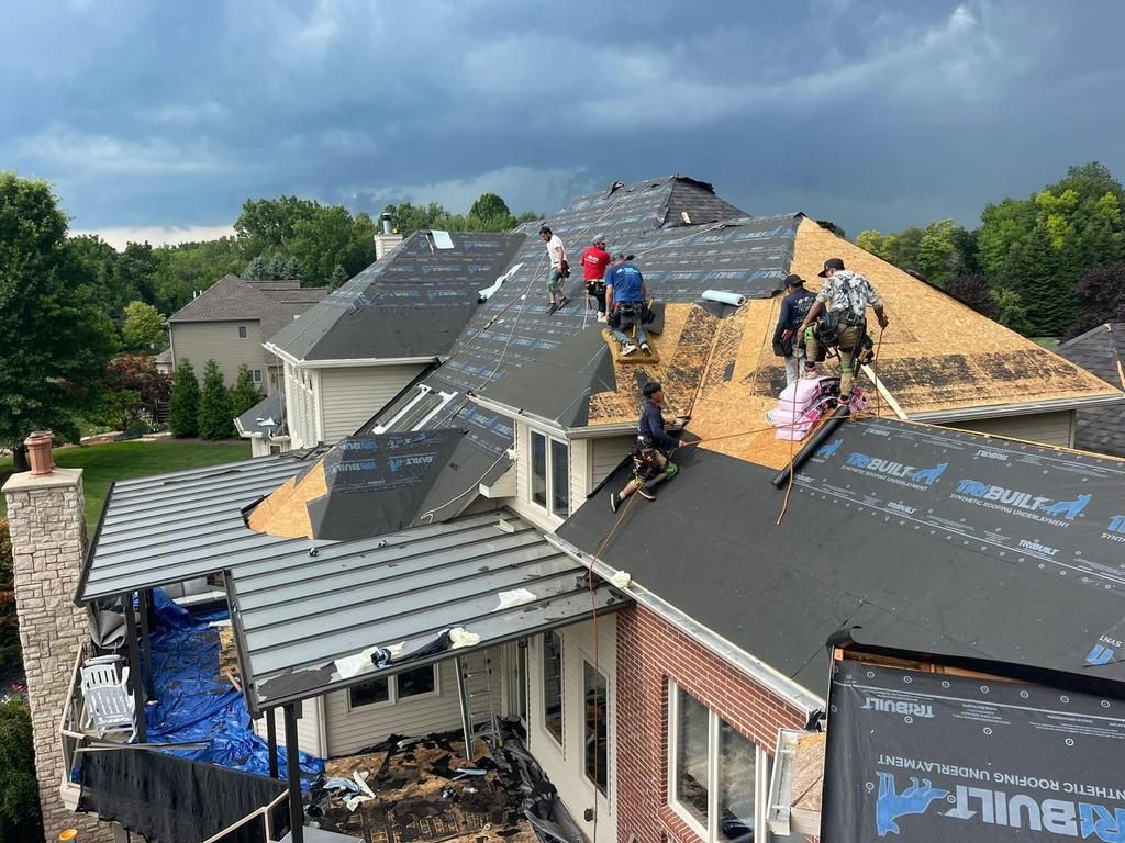 Several workers on a house roof undergoing repairs, with sections covered in black underlayment and exposed wooden decking.