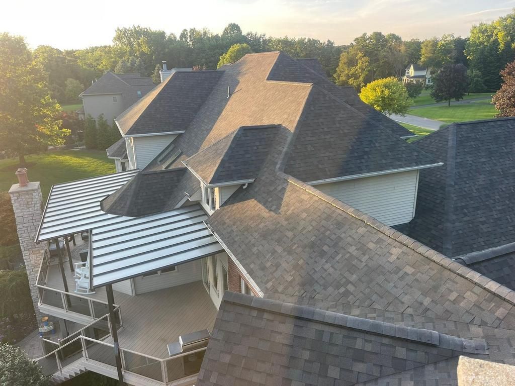 An elevated view of a large residential house with dark shingled roofs, a covered deck, and a stone chimney at sunset.