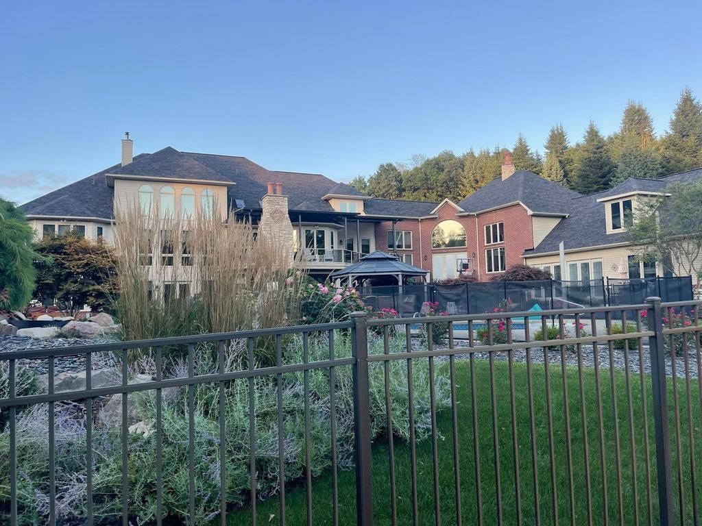 A large multi-story home with a tan exterior and red brick accents, featuring a patio, swimming pool, and metal fence.