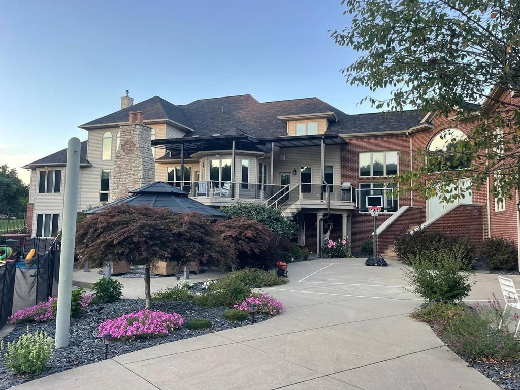 A large, two-story brick and stucco home with a stone chimney, outdoor deck, basketball hoop, and landscaped yard.