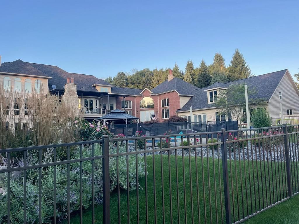 A large suburban house with a swimming pool, backyard, and a metal fence in the foreground under a clear blue sky.