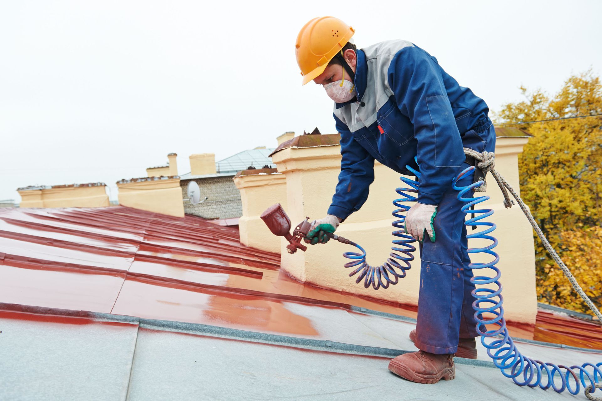 A worker in a helmet and mask spray-paints a red metal roof while connected to a blue coiled air hose.