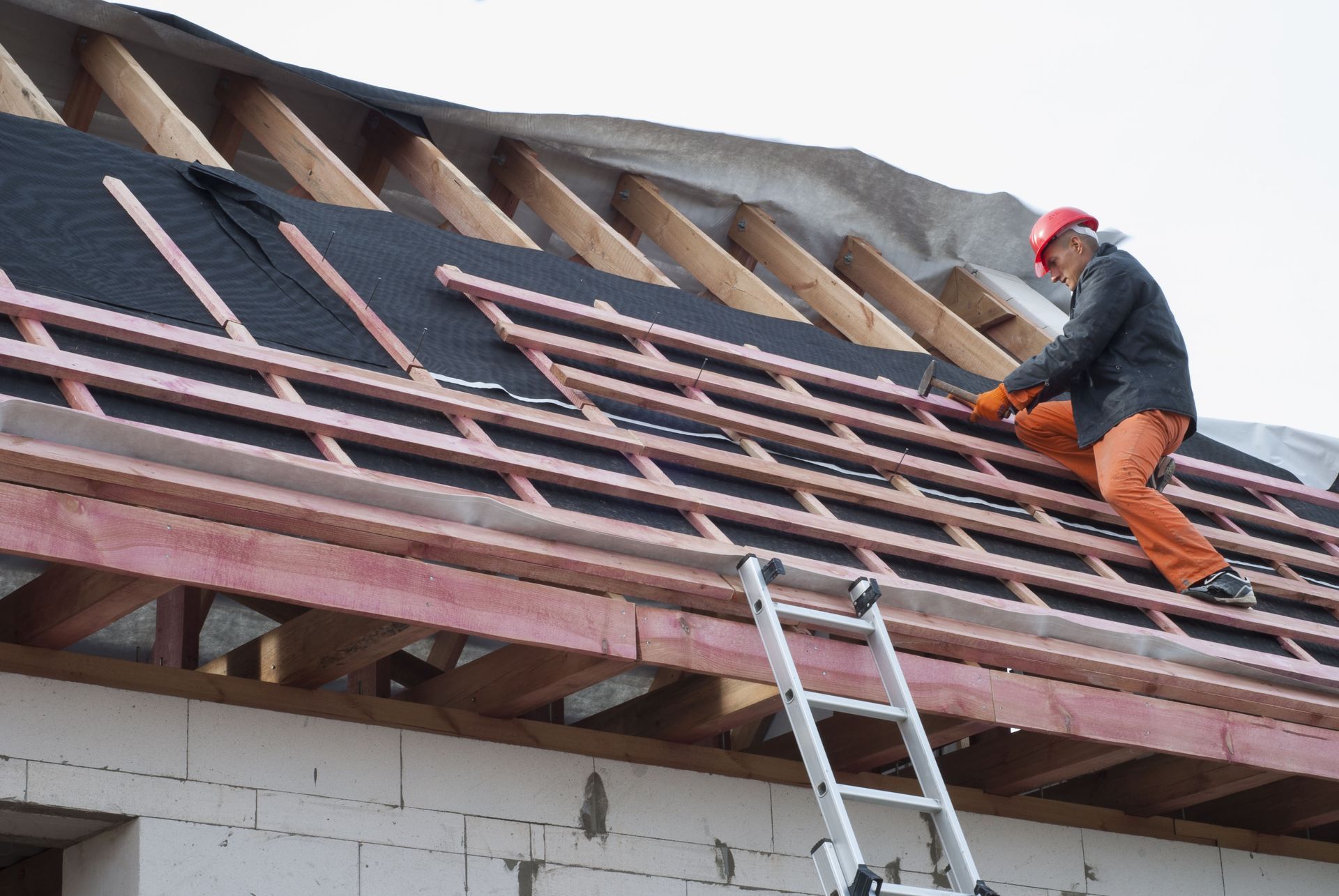 A construction worker in a hard hat and orange pants hammers wooden battens onto a roof framework.