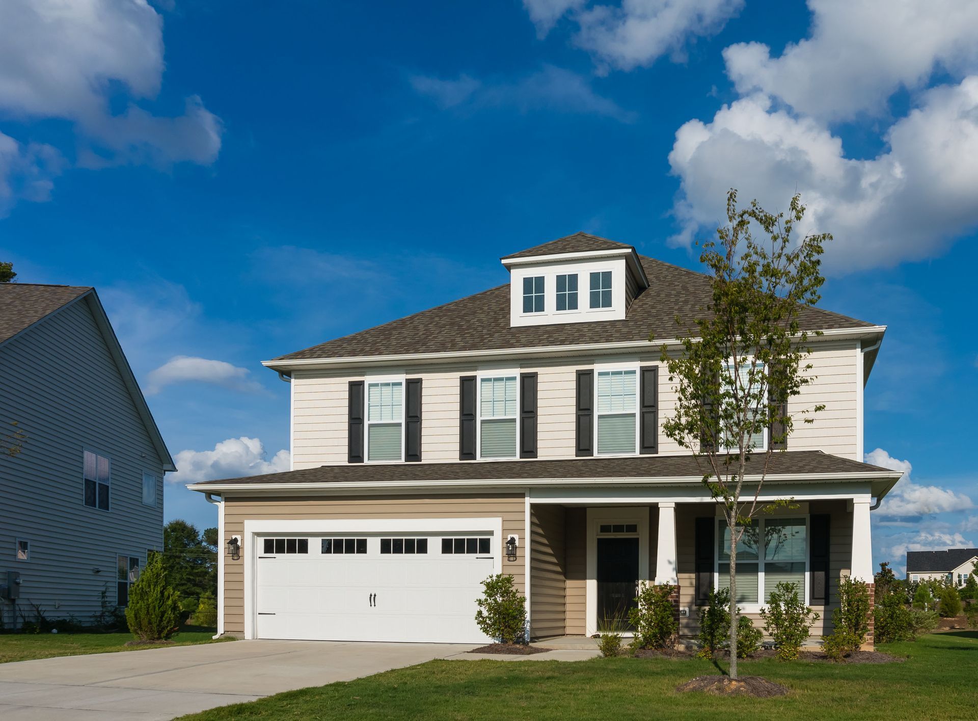 A two-story suburban house with beige siding, a white garage door, black shutters, and a dormer window under a blue sky.