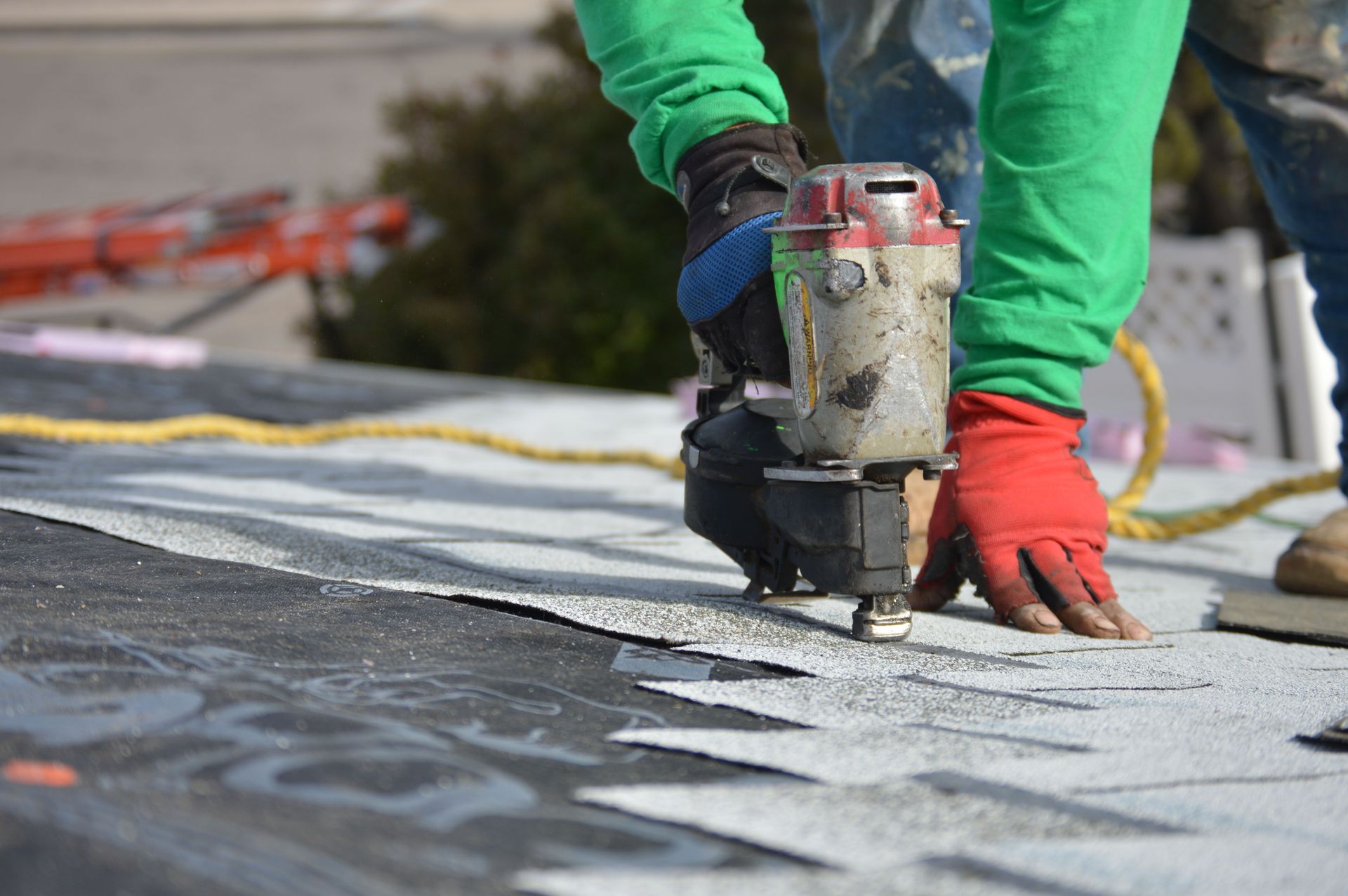 A construction worker in a green long-sleeved shirt uses a pneumatic nail gun to install roofing shingles.