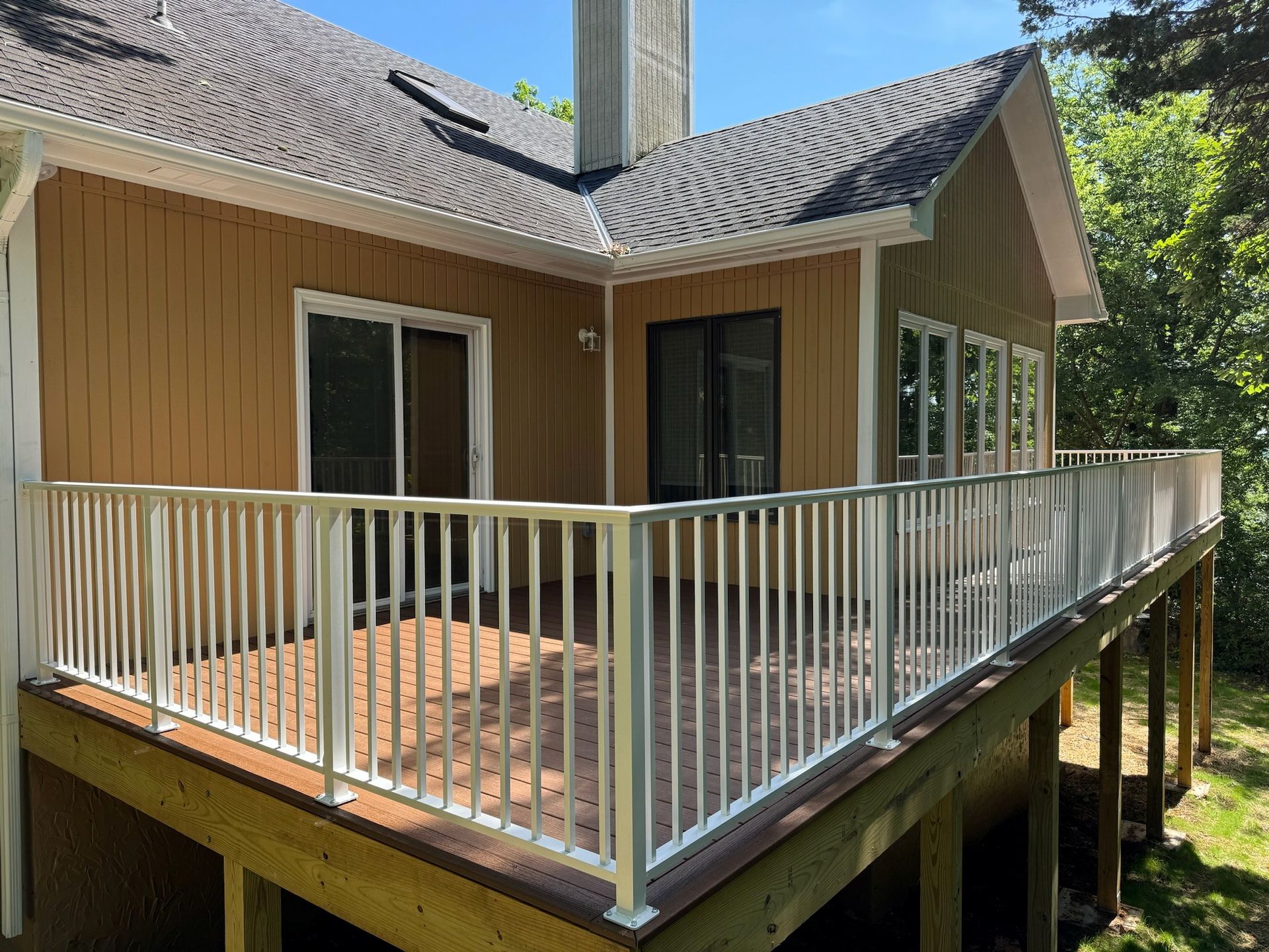 Wraparound wooden deck with white railing on a tan house, viewed from the corner.