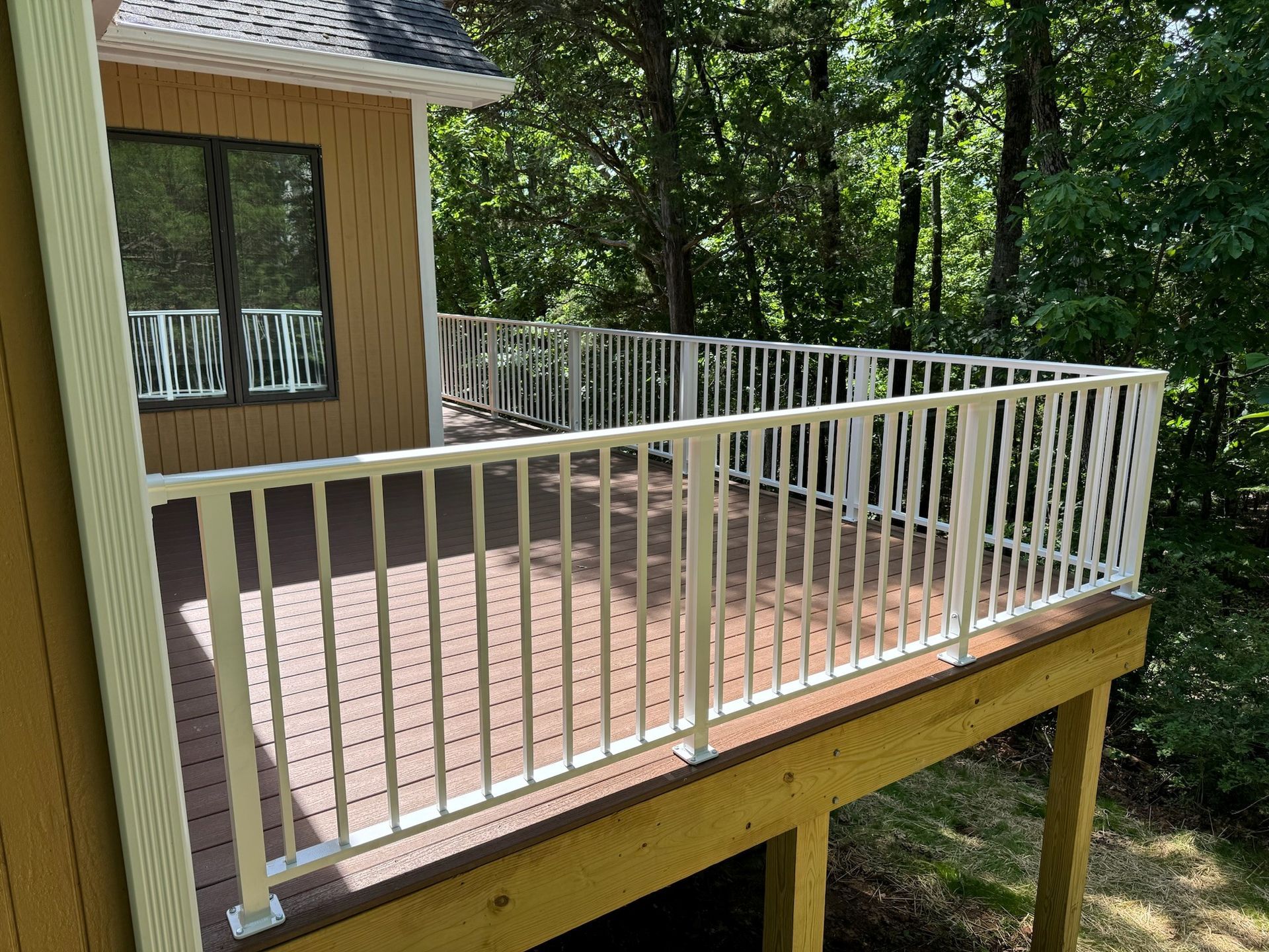 Wooden deck with white railing beside a house, overlooking a wooded area