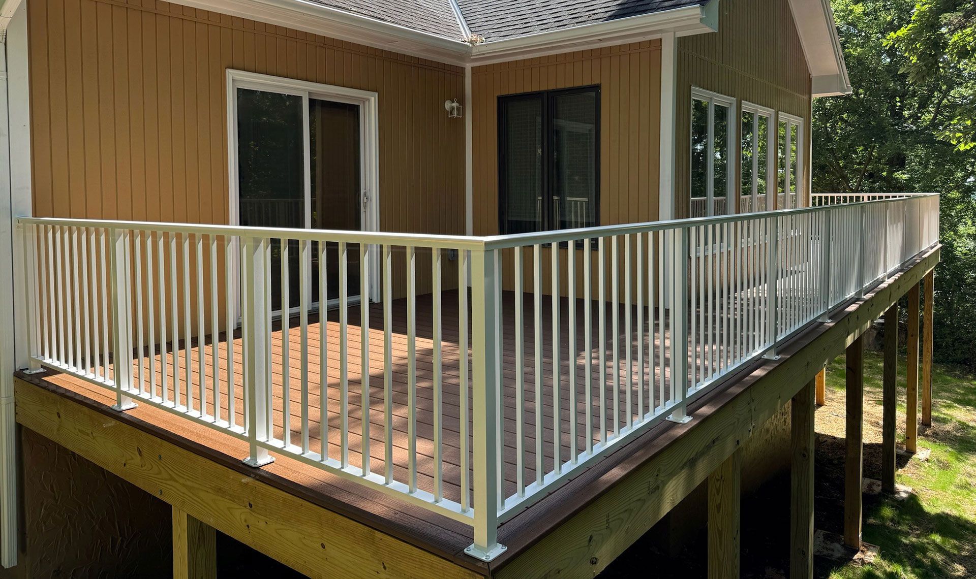 Wooden house deck with white railings and sliding doors, viewed from the side in a wooded yard
