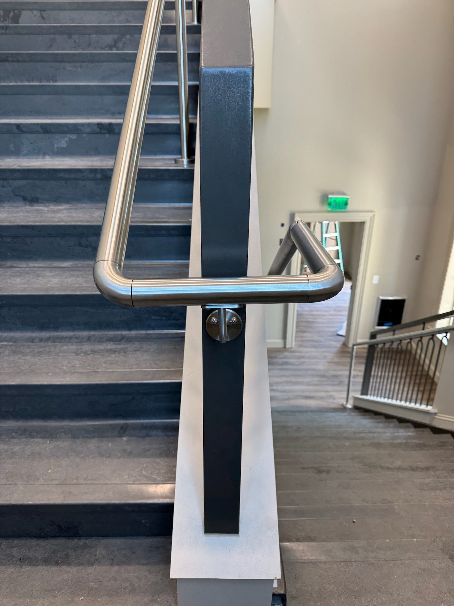 Stairwell with stainless steel handrails, blue steps, and a ramp railing beside a doorway with green exit sign