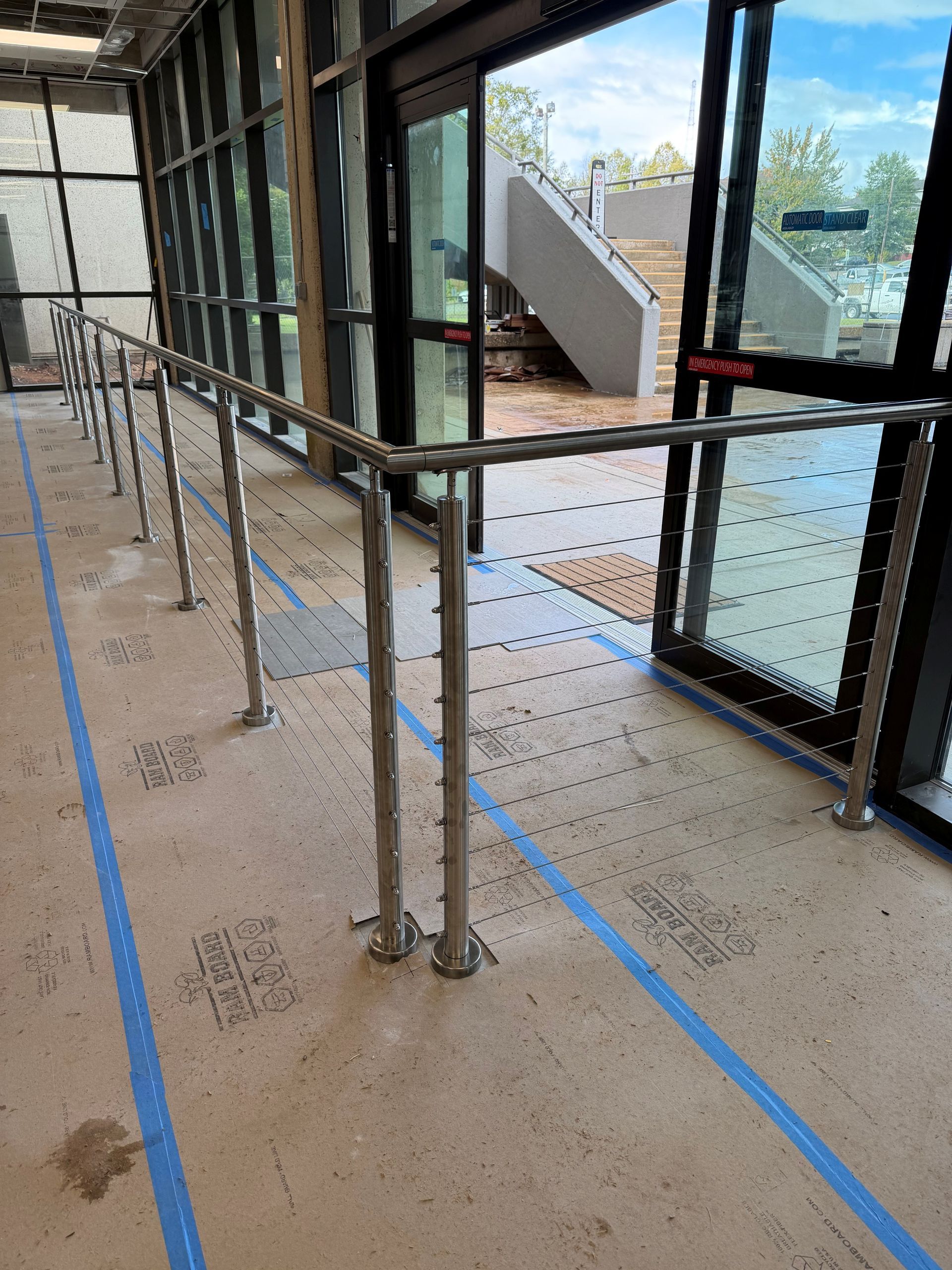 Indoor glass-walled walkway with metal railings, temporary posts, and blue floor markings near a door.