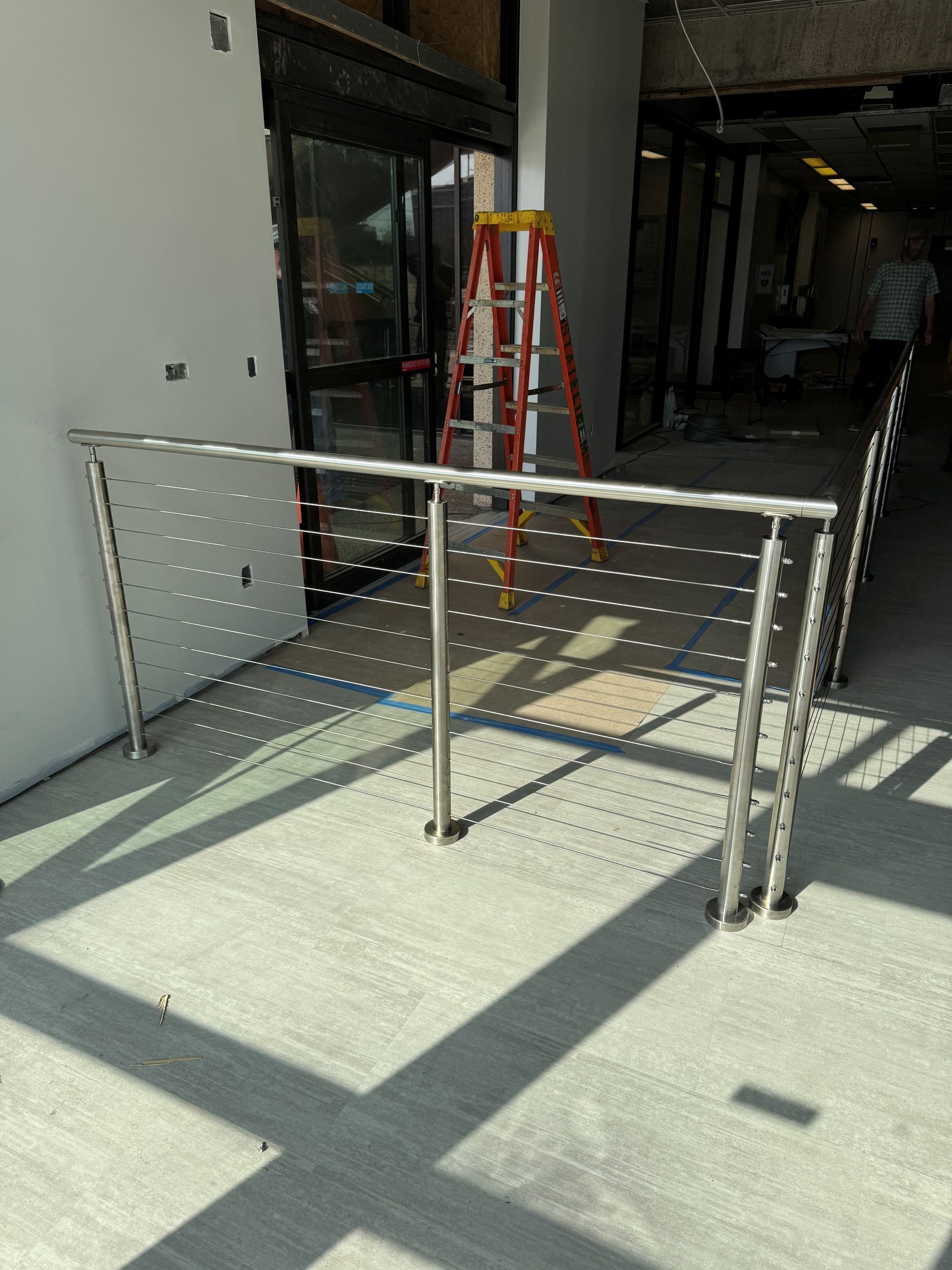 Partially built metal railing in a sunlit industrial hallway, with a red ladder in the background.