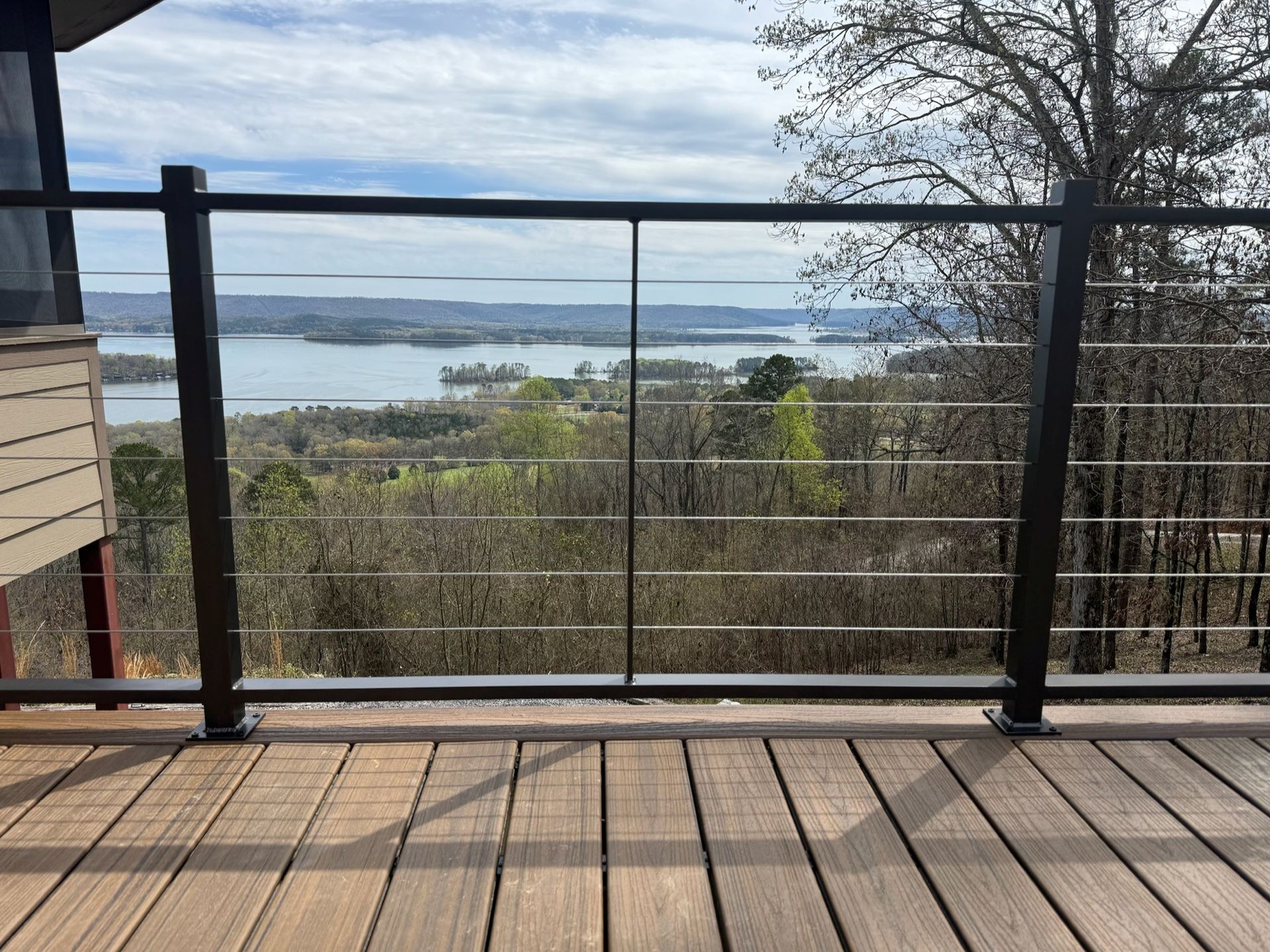 Wooden deck with black railing overlooking trees, water, and distant land under a cloudy sky